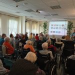 Fotografia przedstawiająca siedzących seniorów oraz śpiewającego do mikrofonu i grającego na gitarze mężczyznę.