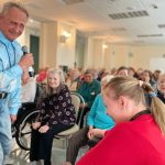 Fotografia przedstawia mężczyznę z mikrofonem, siedzącą obok kobietę. W tle siedzą seniorzy.